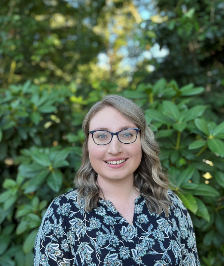 A white woman with brown shoulder length hair and glasses smiling. She is wearing a blue paisley dress and is standing in front of a green bush with trees and sky in the background. 